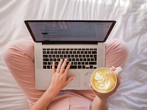 laptop on a bed, hands hold coffee and type at the same time; Top SaaS Blogs for 2022 concept